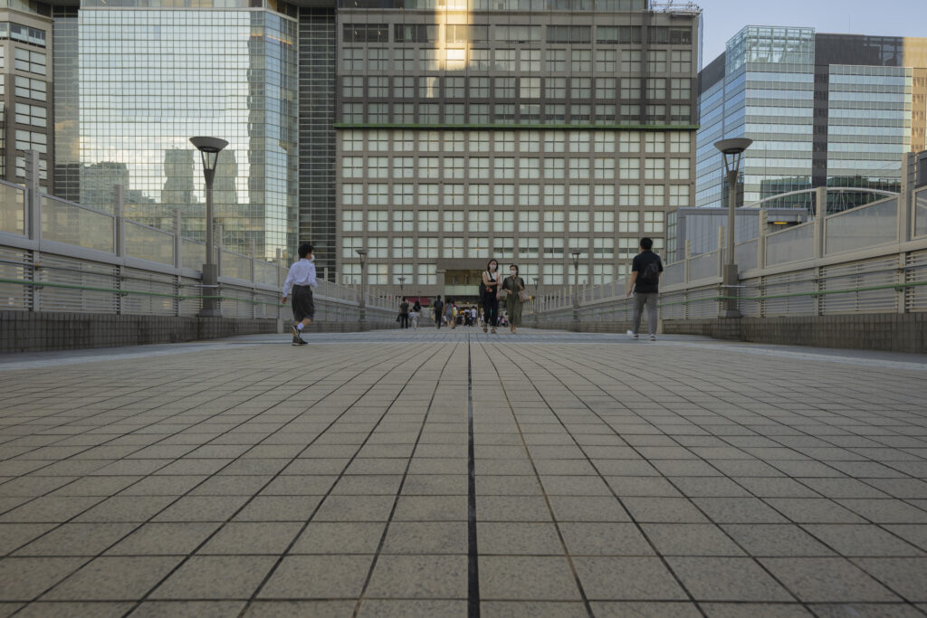 People walking on a modern tiled pedestrian bridge surrounded by tall glass commercial buildings in a busy urban hub.