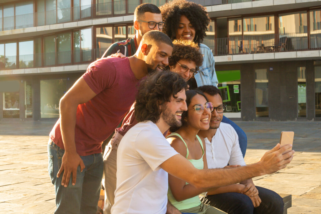 A diverse group of young friends smiling and laughing together while taking a selfie on a smartphone. They are outdoors in a sunny plaza with a modern building in the background.