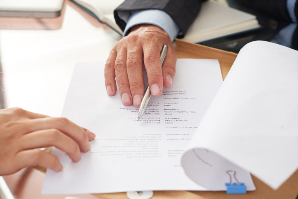 A close up view of a person in a professional suit holding a silver pen while reviewing a printed document on a desk. Another person's hand is visible on the edge of the paper during a formal meeting or review process.