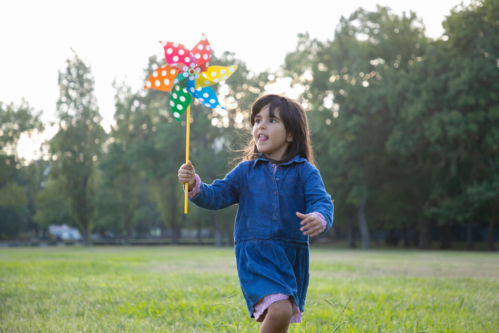A young girl in a blue dress runs through a green grassy park while holding a colorful polka dot pinwheel. The background shows soft sunlight filtering through lush green trees.