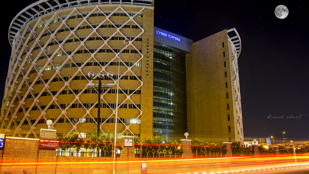 photograph of the iconic cylindrical Cyber Towers building in Hyderabad at dusk, with light trails from fast-moving traffic on the flyover in the foreground.