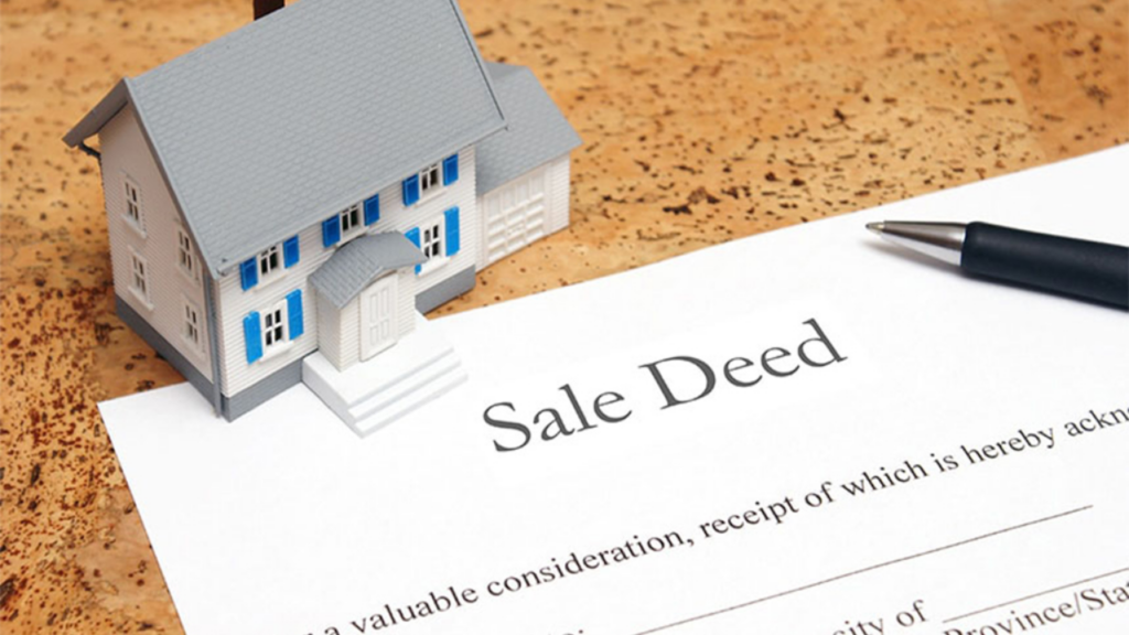 A small model house with blue shutters sits on top of a legal "Sale Deed" document. A black pen rests on the side of the paper.