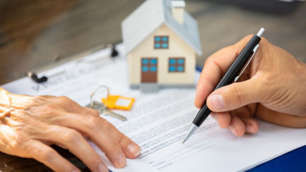 A close up showing hands signing a legal real estate document with a black pen. A model house and a set of keys with a yellow keychain are visible on the desk.