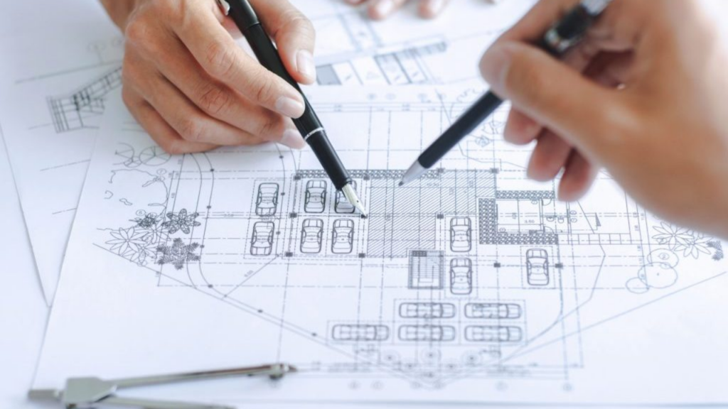 A top-down view of two people in business attire working at a wooden desk. On the table are two small house models, various documents, a pen, and a smartphone displaying a calculator app.