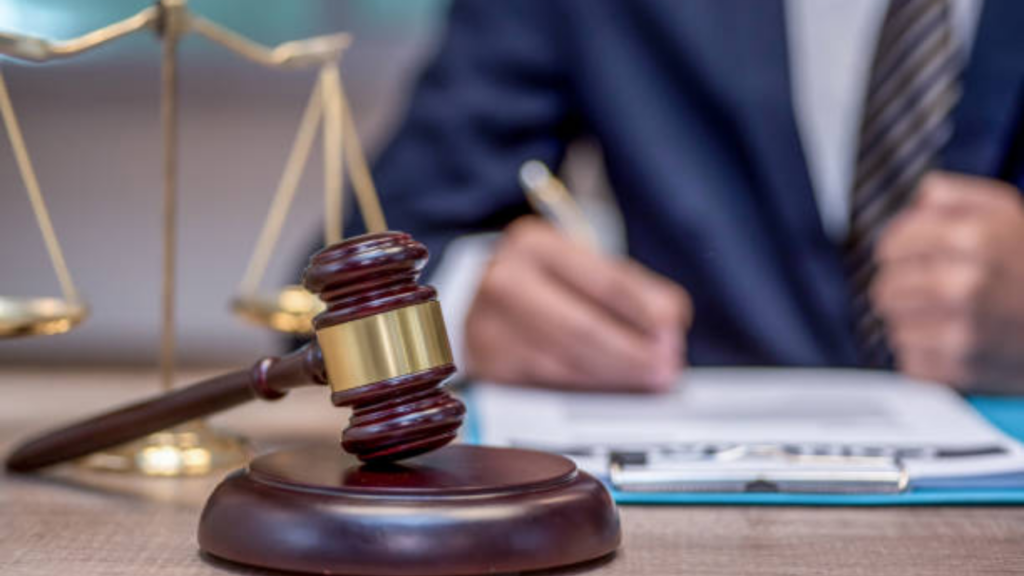 A wooden judge's gavel and golden scales of justice sit on a desk. In the blurred background, a professional in a suit is writing on a clipboard.