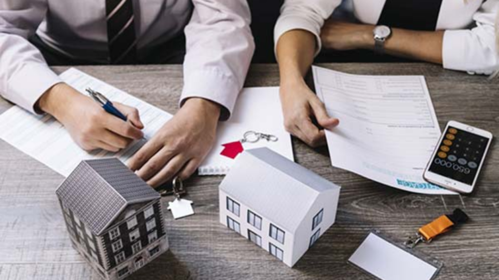 A close-up shot of two people collaborating over a printed blueprint. Both are holding black pens and pointing toward a specific section of a site plan that includes car parking layouts and building footprints.